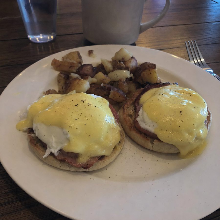 A plate of eggs Benedict shot in dim warm cafe light, then balanced and brightened while the poached eggs, hollandaise, and side potatoes stay unchanged on the same table