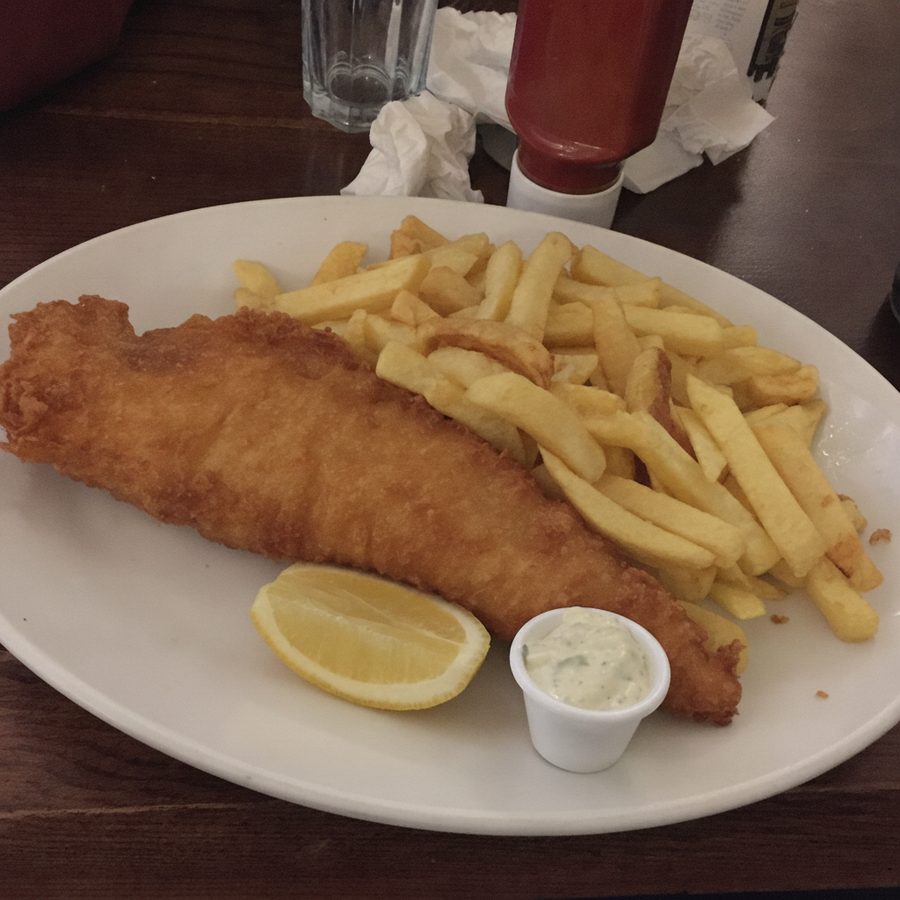 A plate of fish and chips shot in dim pub light on a cluttered table, then brightened and cleaned up while the battered fish, chips, lemon wedge, and tartar sauce stay unchanged