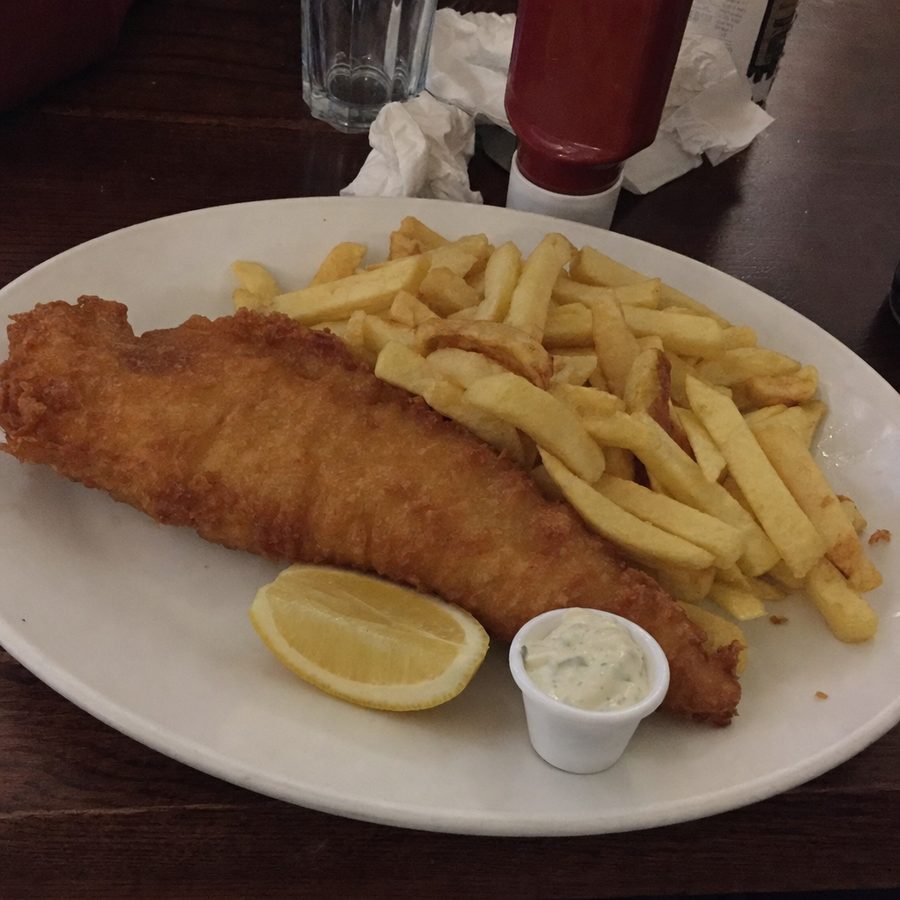 A plate of fish and chips shot in dim pub light on a cluttered table, then brightened and cleaned up while the battered fish, chips, lemon wedge, and tartar sauce stay unchanged