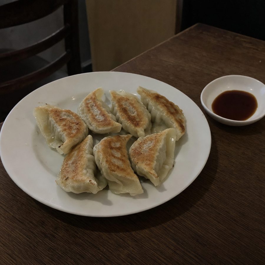 An underexposed phone photo of pan-fried dumplings edited into a brighter menu-ready image with sesame, scallion, and a dipping dish on a warm restaurant table
