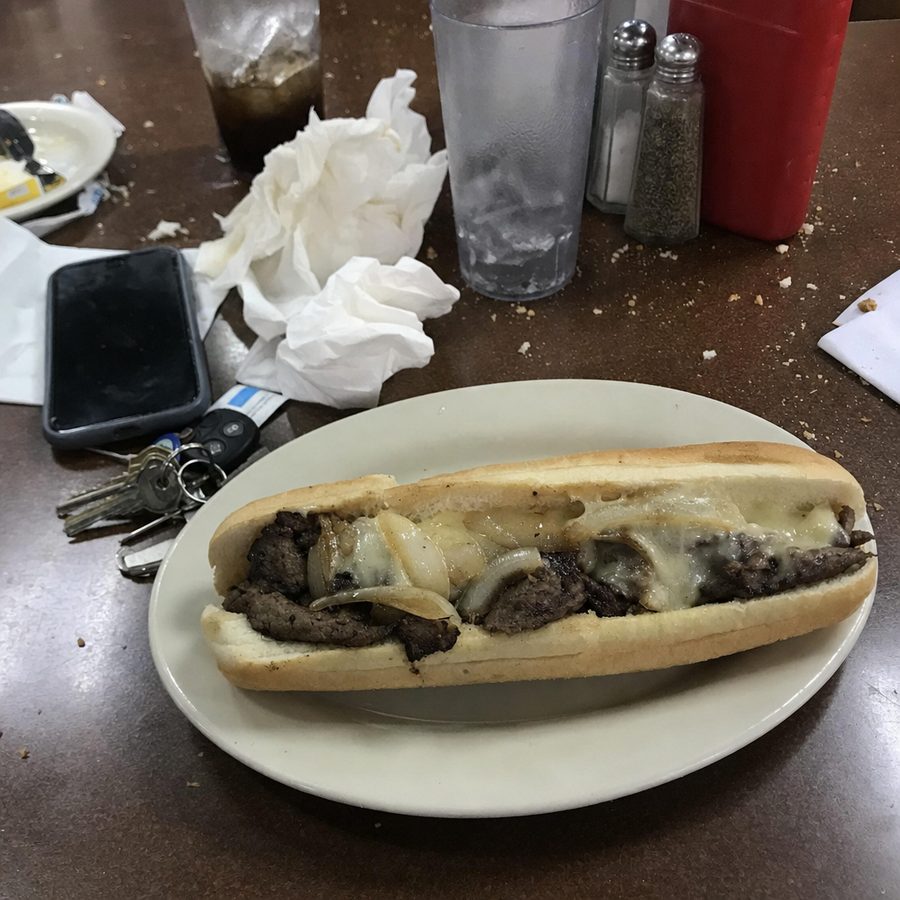 Cluttered, off-center phone photo of a philly cheesesteak on a messy diner table, the kind of shot DoorDash rejects