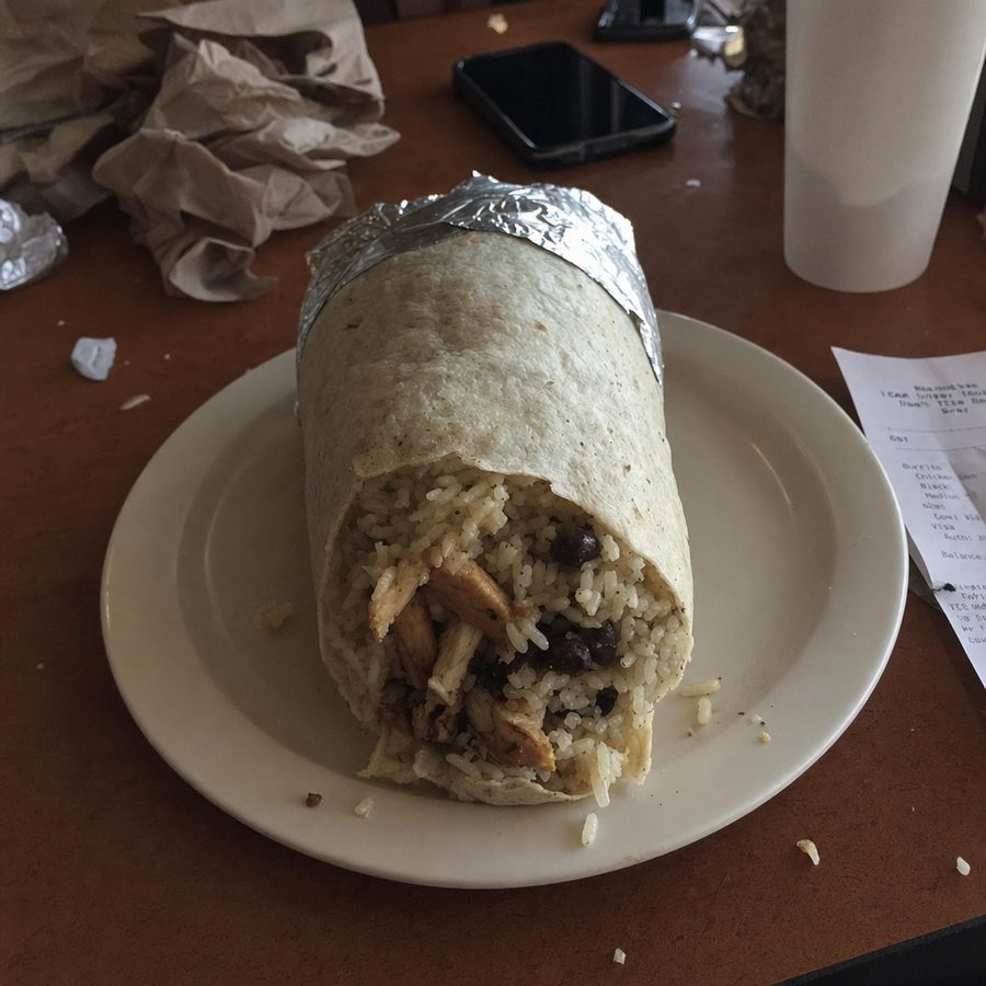Cluttered phone photo of a chicken burrito shot from an awkward angle on a messy table, the kind Uber Eats rejects